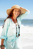 A brunette woman wearing a hat, a necklace and a light-blue beach dress by the sea