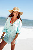 A brunette woman wearing a hat and a light-blue beach dress by the sea