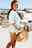 A brunette woman with a hat wearing a light-blue beach dress by the sea