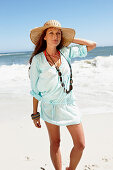 A brunette woman wearing a hat, a necklace and a light-blue beach dress by the sea