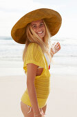 Blonde Frau mit Hut in gelbem Stricktop und Bikini am Strand