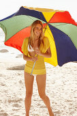 A blonde woman on a beach with a parasol wearing a yellow knitted top and a bikini