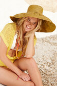 Blonde Frau mit Hut in gelbem Stricktop und Bikini am Strand