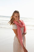 A blonde woman by the sea wearing a salmon-pink scarf and a white dress