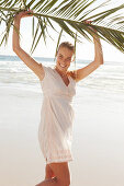 A blonde woman by the sea holding a palm frond wearing a white dress