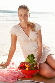 A blonde woman by the sea with a bowl of fruit and vegetables wearing a white dress