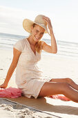A blonde woman on a beach mat by the sea wearing a white dress and a hat