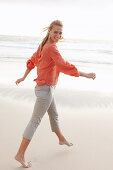 A young blonde woman wearing an orange blouse and light trousers by the sea