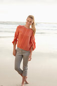 A young blonde woman wearing an orange blouse and light trousers by the sea