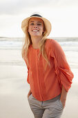 Junge blonde Frau mit Hut in orangefarbener Bluse und heller Hose am Meer