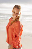 Junge blonde Frau in orangefarbener Bluse und heller Hose am Meer