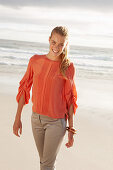 A young blonde woman wearing an orange blouse and light trousers by the sea