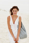 A brunette woman by the sea with a bag wearing a white summer dress