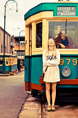 A young couple in front of and inside a tram, her wearing a summer dress and him wearing a shirt and a jacket