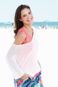Junge Frau in lachsfarbenem Top, weißem Pulli und bunter Hose am Strand