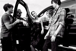 A young woman wearing a black party dress and young men wearing casual outfits standing by a car (black-and-white shot)