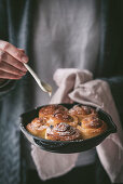 Small spoon adding sweet sauce to delicious fresh cinnamon rolls on frying pan