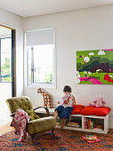 Girl with book sits on bench, green upholstered armchair in foreground