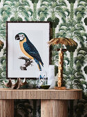 Lamp in the shape of a palm tree and picture of a parrot in front of wallpaper with palm tree motifs