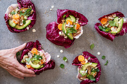 Man’s hand taking one of several healthy, eatable radicchio bowls with couscous, avocado, dates. carrots, cucumber, herbs and sesam seeds