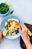 Woman eating Turkish eggs with some toasted bread, baby spinach in a small blue bowl accompanies