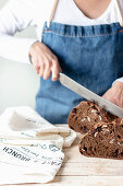 Hands of woman in apron holding long steel knife and cutting rye bread with raisins and nuts