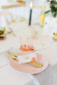 Festlich gedeckter Tisch mit Namenskärtchen und Goldbesteck auf zartrosafarbenem Teller