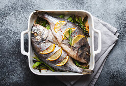 Baked fish dorado with green asparagus in white ceramic baking pan on gray rustic concrete background