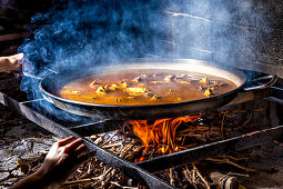 Hands holding big iron pan with boiling broth for cooking paella over open fire with wood