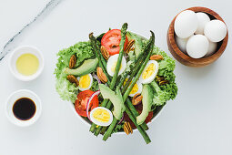 Served bowl of salad with asparagus eggs avocados tomatoes walnuts and greenery on table with condiments sauces and eggs