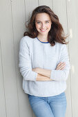 A young brunette woman standing in front of a wooden wall wearing a light knitted jumper and jeans