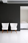 Two white retro armchairs against black wall with view into bedroom