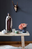 Flowers and Madonna figurine on wooden bench in front of hunting trophy on dark wall