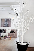 White-painted tree decorated with baubles in front of picture of Buddha above sofa