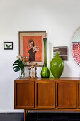 Green glass vases, candlesticks and plants on retro sideboard below artworks on wall