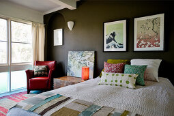 Bett mit Kissen und Stoffmuster, Nachtkasten und roter Ledersessel, Bilder an dunkler Wand im Schlafzimmer