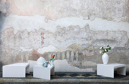 Vases and hand-painted china bird on small, white, designer tables in front of artistically distressed wall