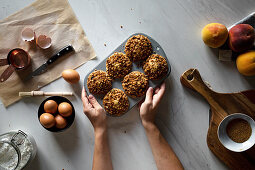 From above crop person holding baking dish with homemade cupcakes on wooden table with arranged eggs with peaches and flour