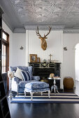 Blue-and-white armchair and footstool with toile de jouy upholstery in front of fireplace below hunting trophy