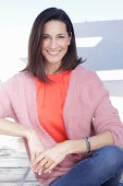 A mature brunette woman wearing a red blouse, a wrap-around jumper and jeans