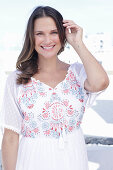 A long-haired woman wearing a white summer dress with colourful embroidery detail