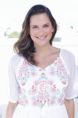 A long-haired woman wearing a white summer dress with colourful embroidery detail