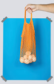 Hand carrying orange net sack with fresh eggs against blue rectangle during zero waste shopping