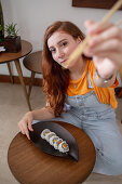 Young redhead woman in casual clothes looking at camera using chopsticks while sitting at table and eating sushi at home
