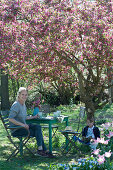 Seating group under the crab apple tree 'Paul Hauber', woman with coffee cup, boy sitting and reading, dog Zula