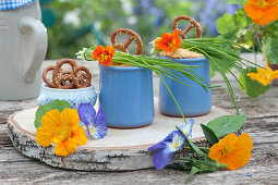 Mug with obatzder, pretzels, chives, flowers of nasturtium and horned violets on a wooden board made of birch wood