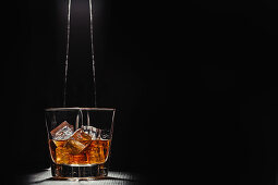 Person filling transparent glass with bourbon on wooden table in rays against black background