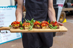 Person serviert belegte Brote mit Wurst, Tomaten und Rucola auf Holzbrett