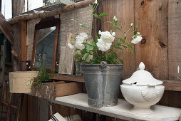 Suppenterrine und Zinkeimer mit weißen Rosen auf Holzablage