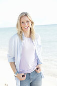 A blonde woman by the sea wearing a purple t-shirt and a blue-and-white striped shirt